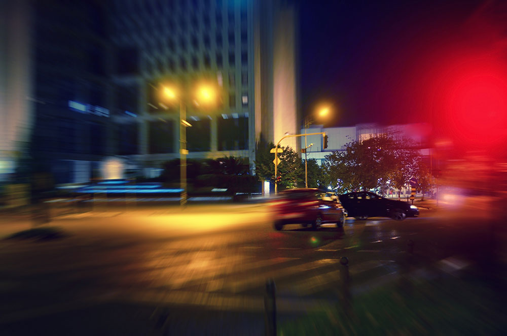 A city intersection at night, with a motion-blurred red vehicle about to run a red light and hit another car.